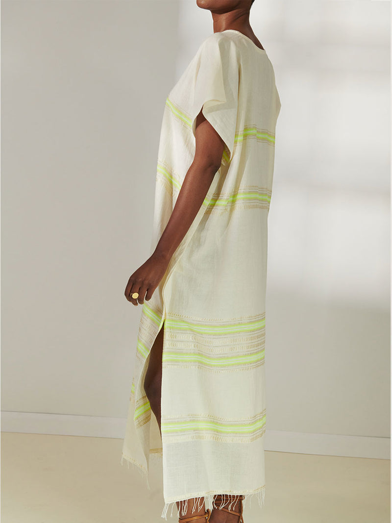 Side View of a Woman Standing Wearing lemlem Dalila Caftan featuring combination of matte and shine natural tibebs and stripes in Vanilla Cream and Lime sorbet colors.