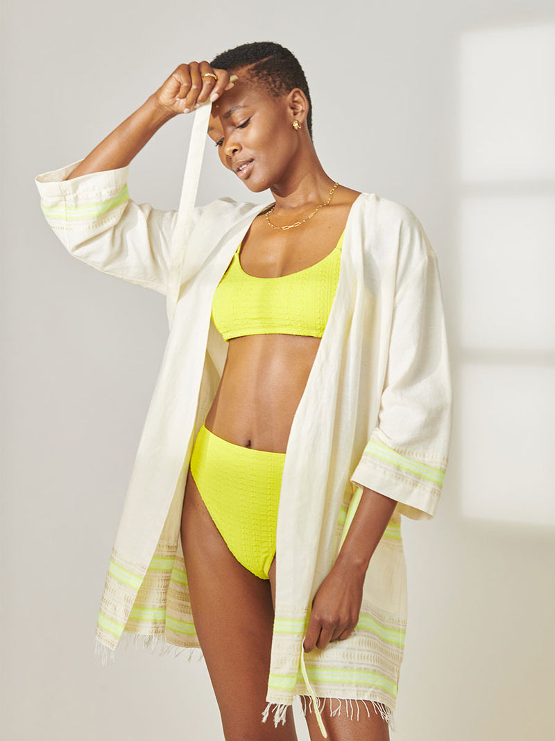 Woman Standing wearing lemlem Imani Short Robe featuring combination of matte and shine natural tibebs and stripes in Vanilla Cream and Lime sorbet colors and Jordanos Citron Scoop Top and High Waist Bottom