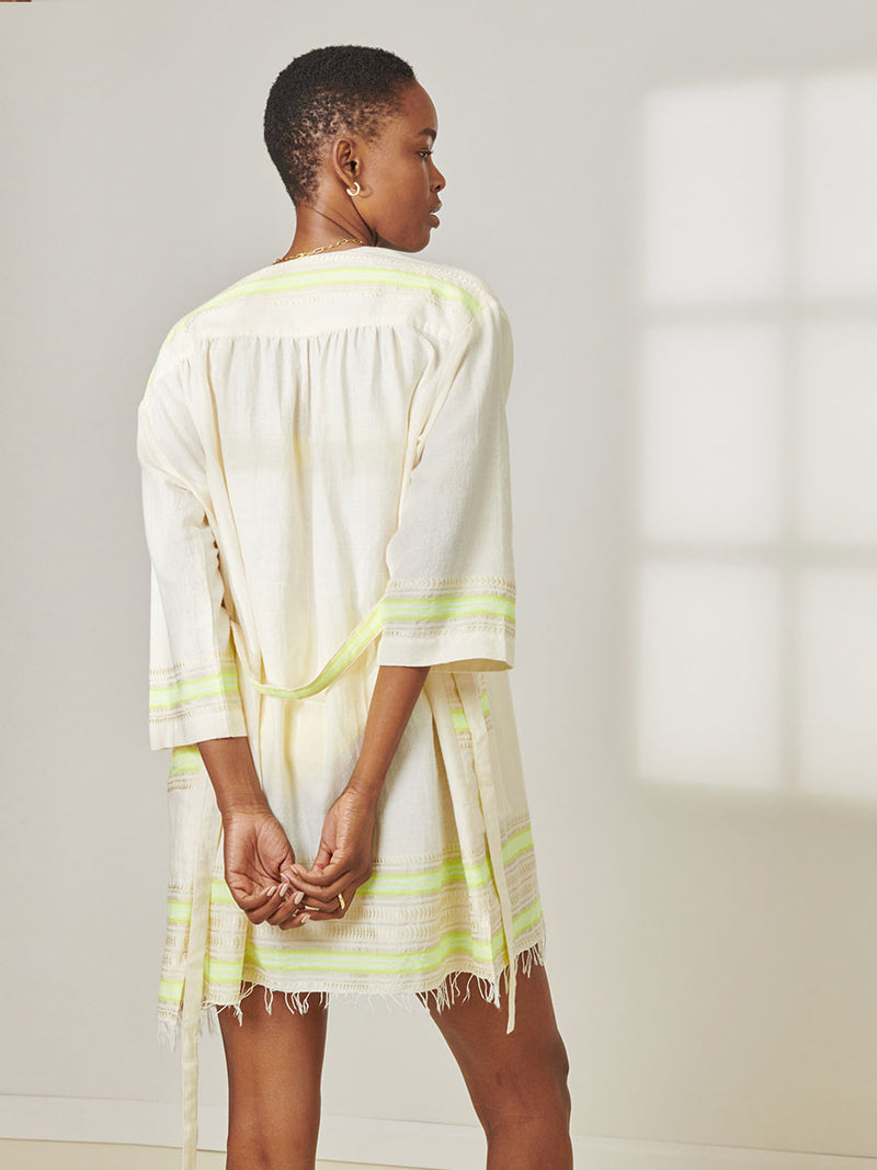 Back View of a Woman Standing wearing lemlem Imani Short Robe featuring combination of matte and shine natural tibebs and stripes in Vanilla Cream and Lime sorbet colors