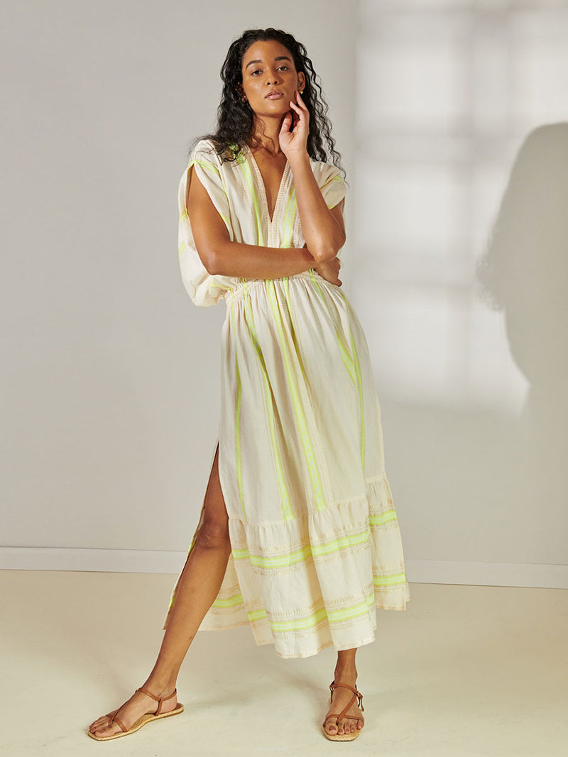 Woman Standing Wearing lemlem Leila Plunge Neck Dress featuring combination of matte and shine natural tibebs and stripes in Vanilla Cream and Lime sorbet colors.