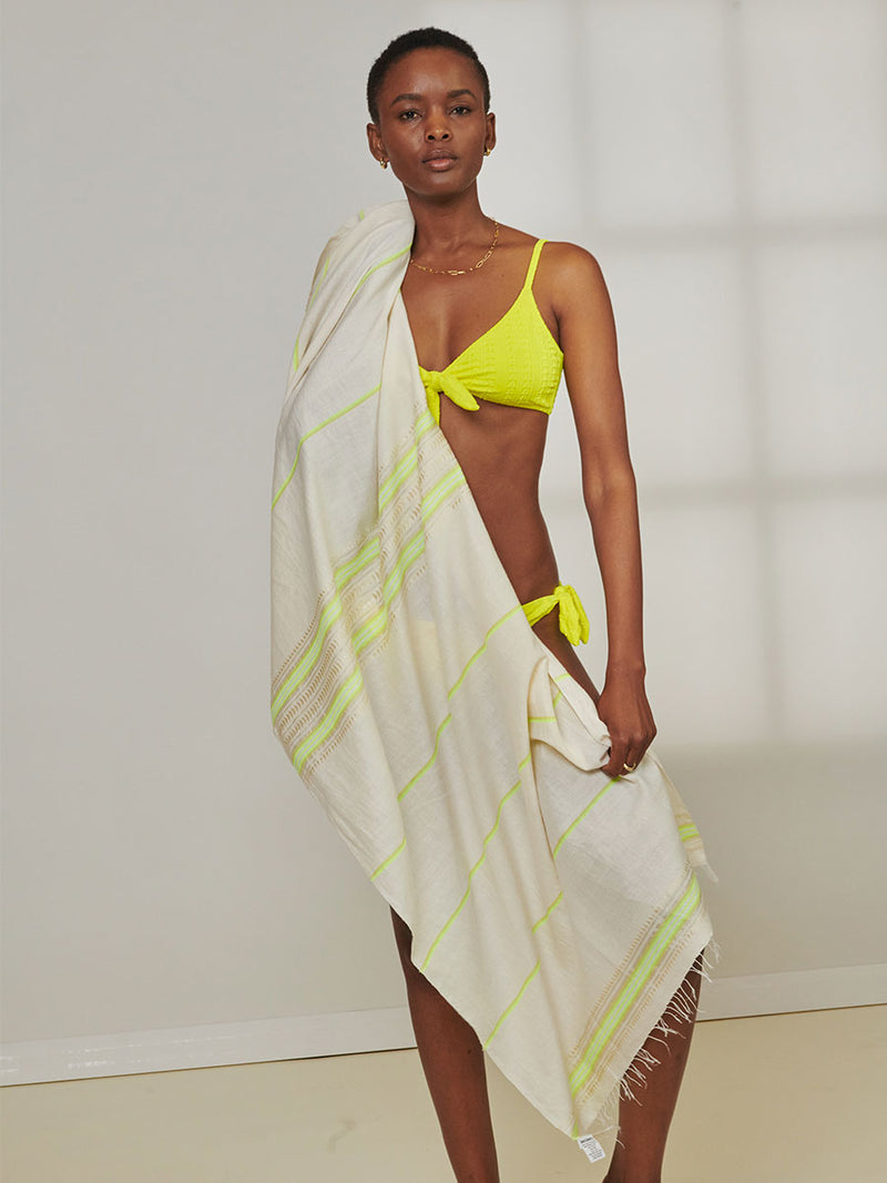 Woman Standing Wearing lemlem Jordanos Citron Tie Front Top and Side Tie Bottom and holding lemlem  Lema Sarong featuring combination of matte and shine natural tibebs and stripes in Vanilla Cream and Lime sorbet colors.