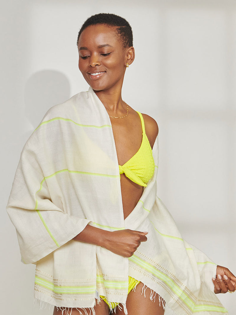 Woman Standing Wearing lemlem Jordanos Citron Tie Front Top and Side Tie Bottom and holding lemlem Lema Sarong featuring combination of matte and shine natural tibebs and stripes in Vanilla Cream and Lime sorbet colors.