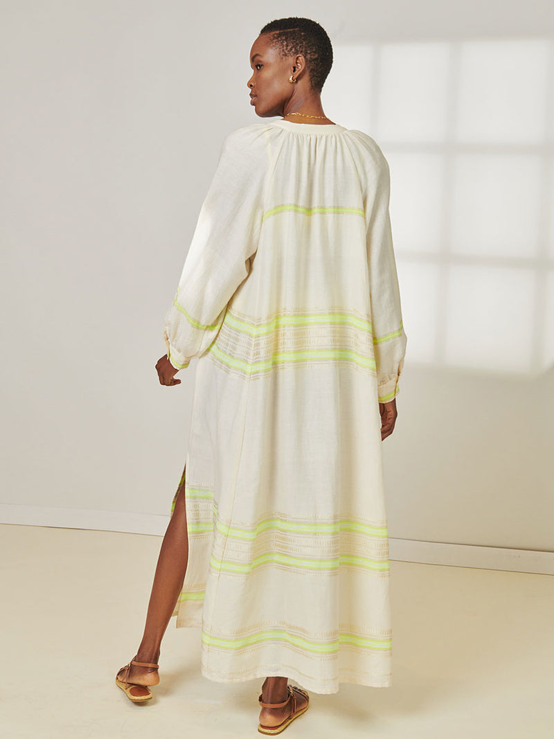 Back View of a Woman Standing Wearing lemlem Makeda Button Up Dress featuring combination of matte and shine natural tibebs and stripes in Vanilla Cream and Lime sorbet colors.