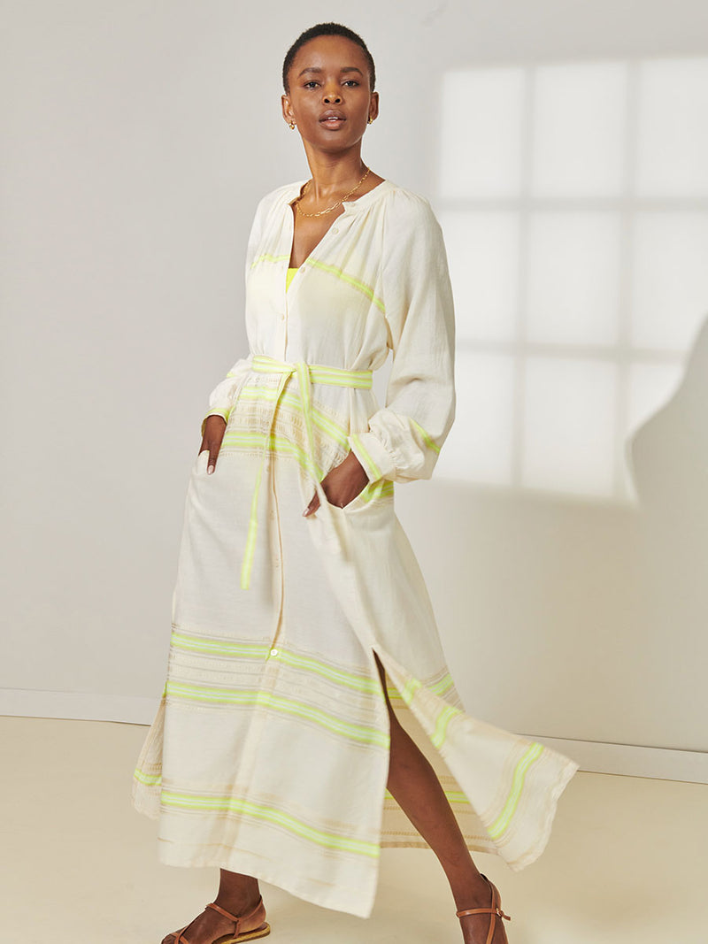 Woman Standing Wearing lemlem Makeda Button Up Dress featuring combination of matte and shine natural tibebs and stripes in Vanilla Cream and Lime sorbet colors.