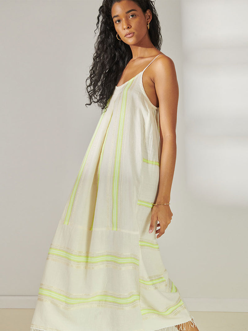 Side View of a Woman Standing Wearing lemlem Nia Slip Dress featuring combination of matte and shine natural tibebs and stripes in Vanilla Cream and Lime sorbet colors.
