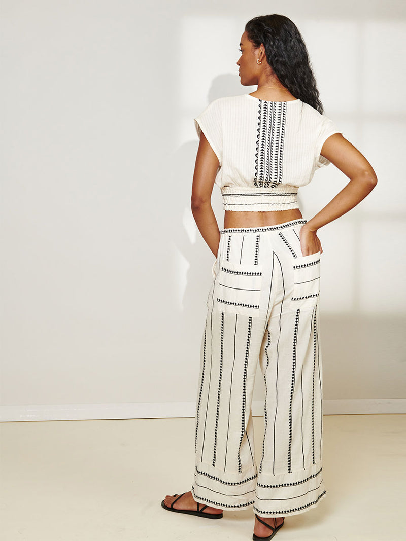 Back View of a Woman Standing Wearing Desta Pants featuring intricate black Tibeb bands on a textured vanilla background and matching Alia Plunge Top