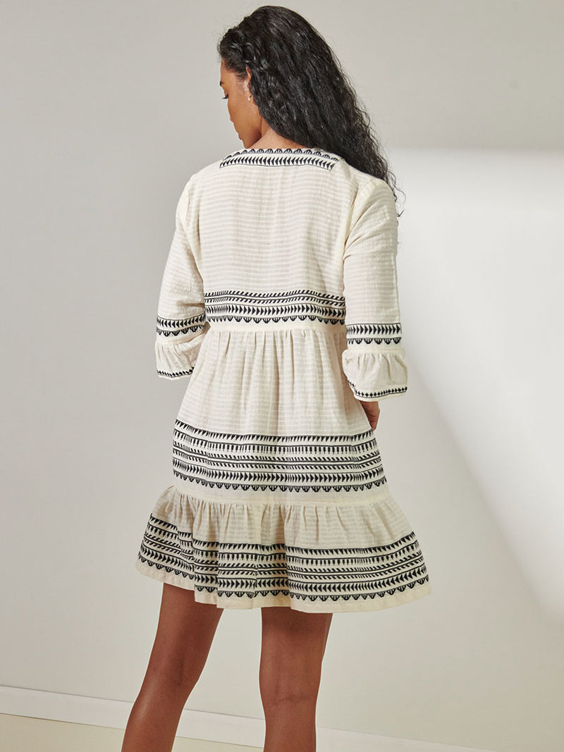 Back View of Woman Standing Wearing lemlem Hanna Flutter Dress featuring intricate black Tibeb bands on a textured vanilla background.