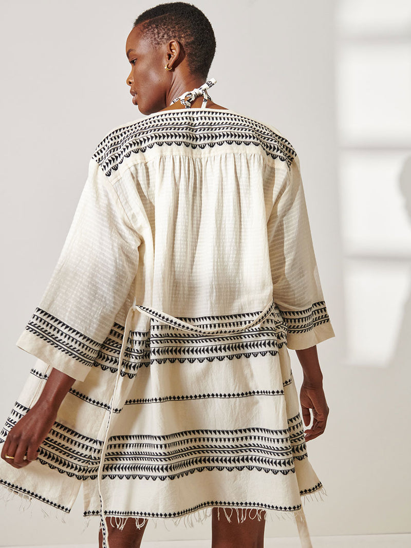 Back View of a Woman Standing Wearing lemlem Imani Short Robe featuring intricate black Tibeb bands on a textured vanilla background