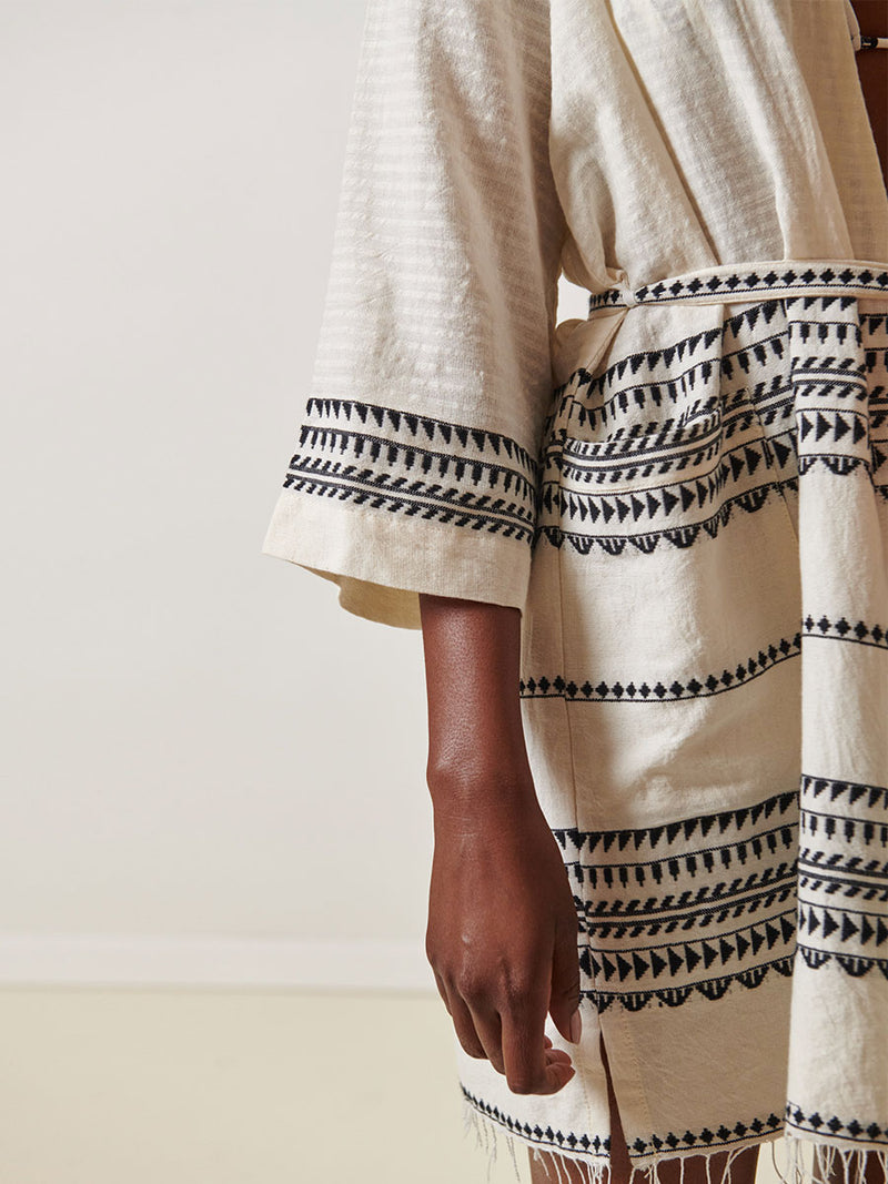 Close Up on a Woman Standing Wearing lemlem Imani Short Robe featuring intricate black Tibeb bands on a textured vanilla background