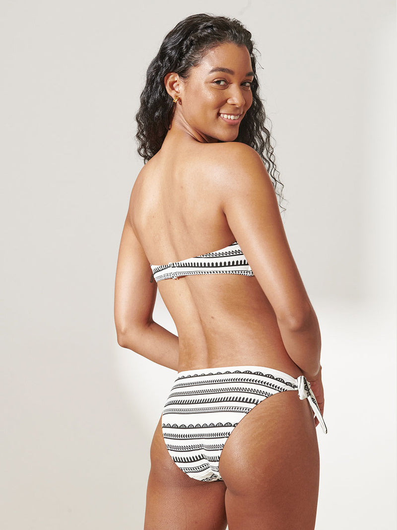 Back View of a Woman Standing Wearing lemlem Lucy Side Tie Bikini Bottom featuring intricate black Tibeb bands on a textured white background and a matching Bandeau Top