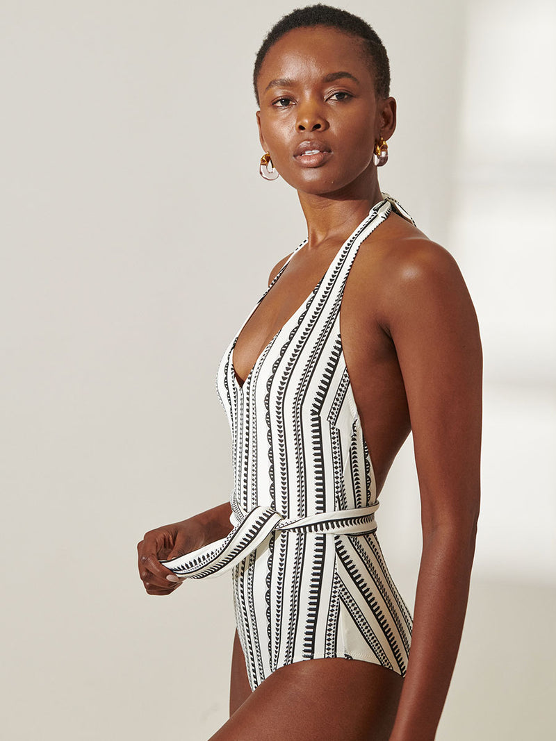 Woman standing wearing lemlem Lidya Deep V One Piece featuring intricate black Tibeb bands on a textured white background.