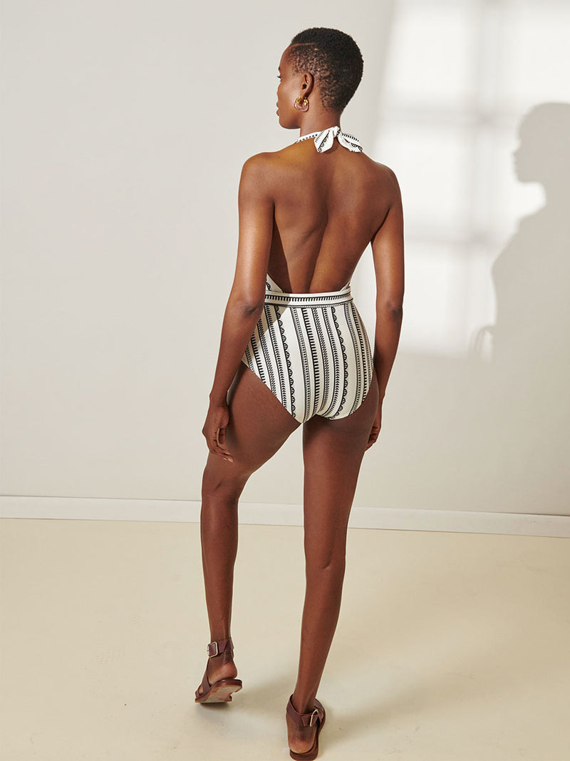 Back View of a Woman standing wearing lemlem Lidya Deep V One Piece featuring intricate black Tibeb bands on a textured white background.