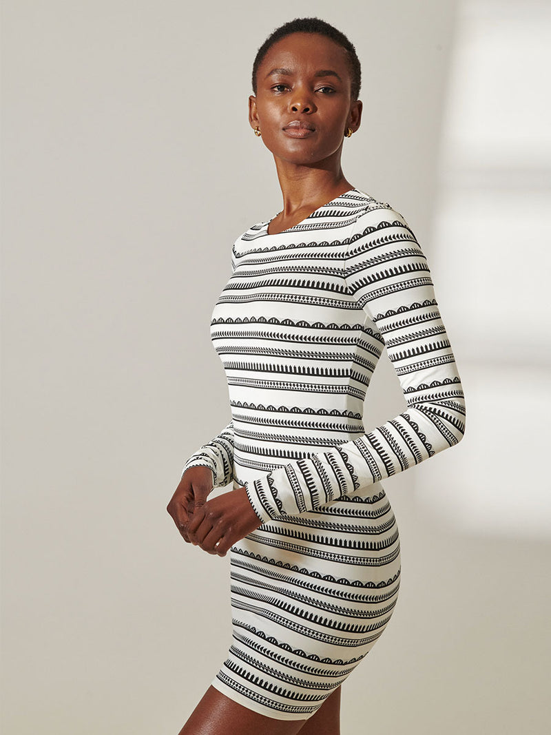 Side View of a woman standing wearing lemlem Nyala Swim Dress featuring intricate black Tibeb bands on a textured white background.