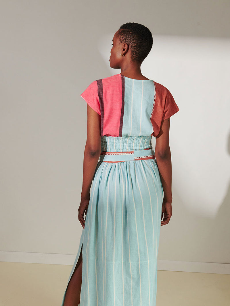 Back View of a Woman Standing Wearing lemlem Alia Plunge Top Featuring all over stripe color block pattern in sky, terracotta, brick and burgundy colors and matching wrap skirt