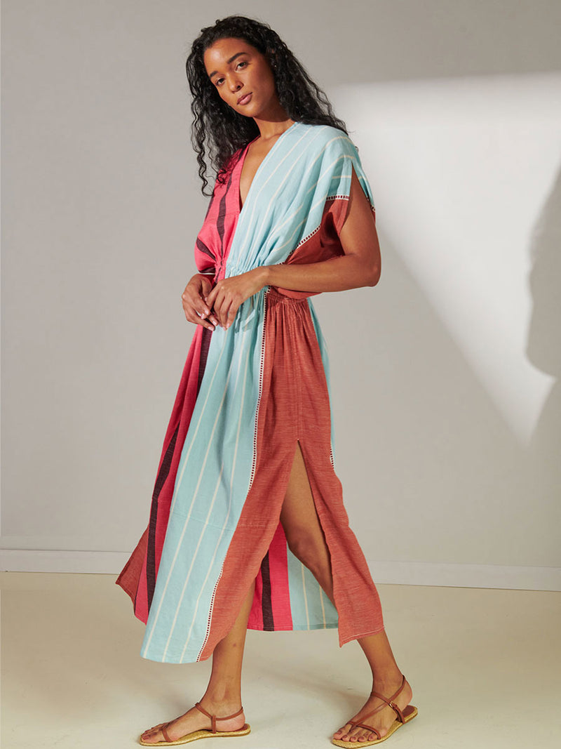 Side View of a Woman Standing Wearing Lelia Plunge Neck Dress Featuring all over stripe color block pattern in sky, terracotta, brick and burgundy colors.