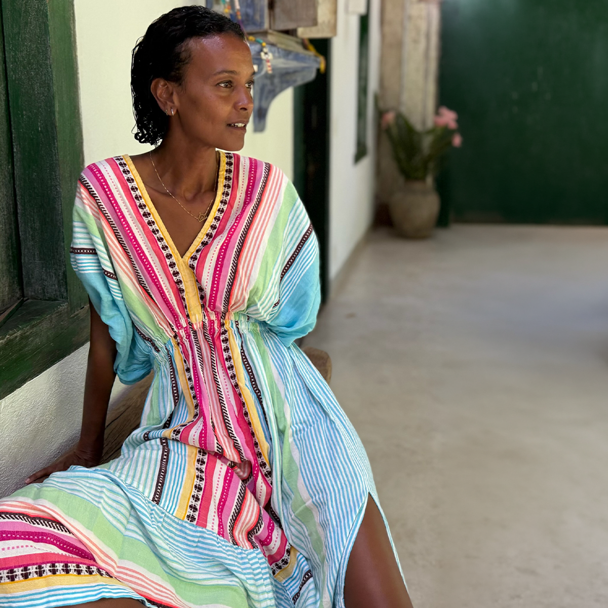 Liya Kebede in a multicolor striped handwoven lemlem dress sitting on a bench indoors.