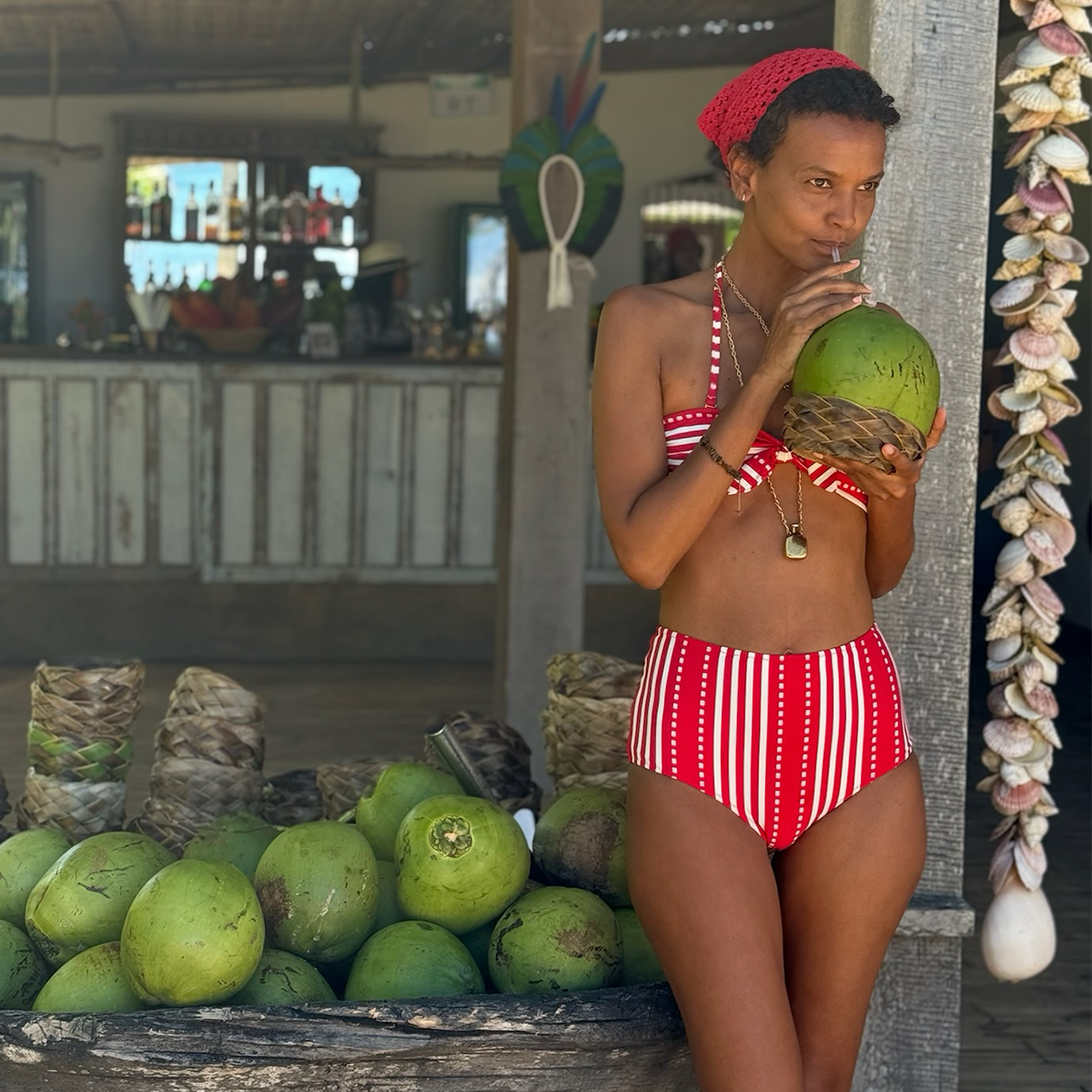 Liya Kebede in a red and white lemlem bikini holding a coconut next to coconuts on display.