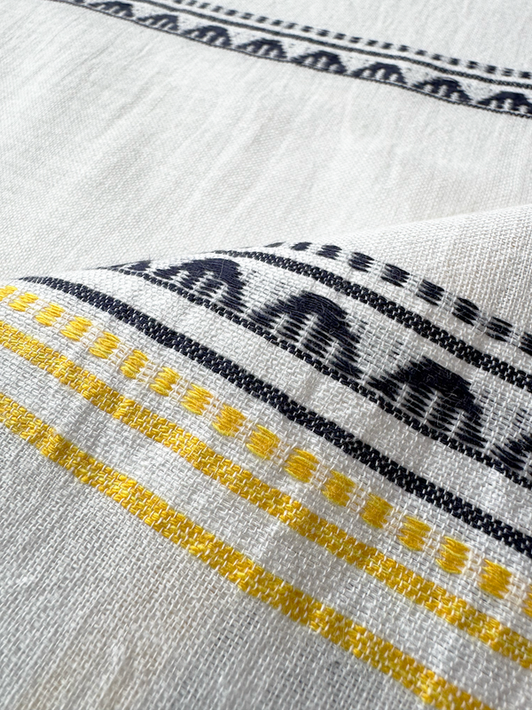 close up on lemlem handwoven sisi fabric featuring an intricate tibeb scallop pattern in navy, accented with yellow on a white background.