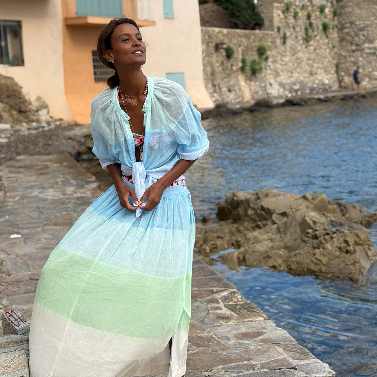Liya Kebede standing in light handwoveb blue and green skirt standing by a waterfront with a stone wall and building in the background.