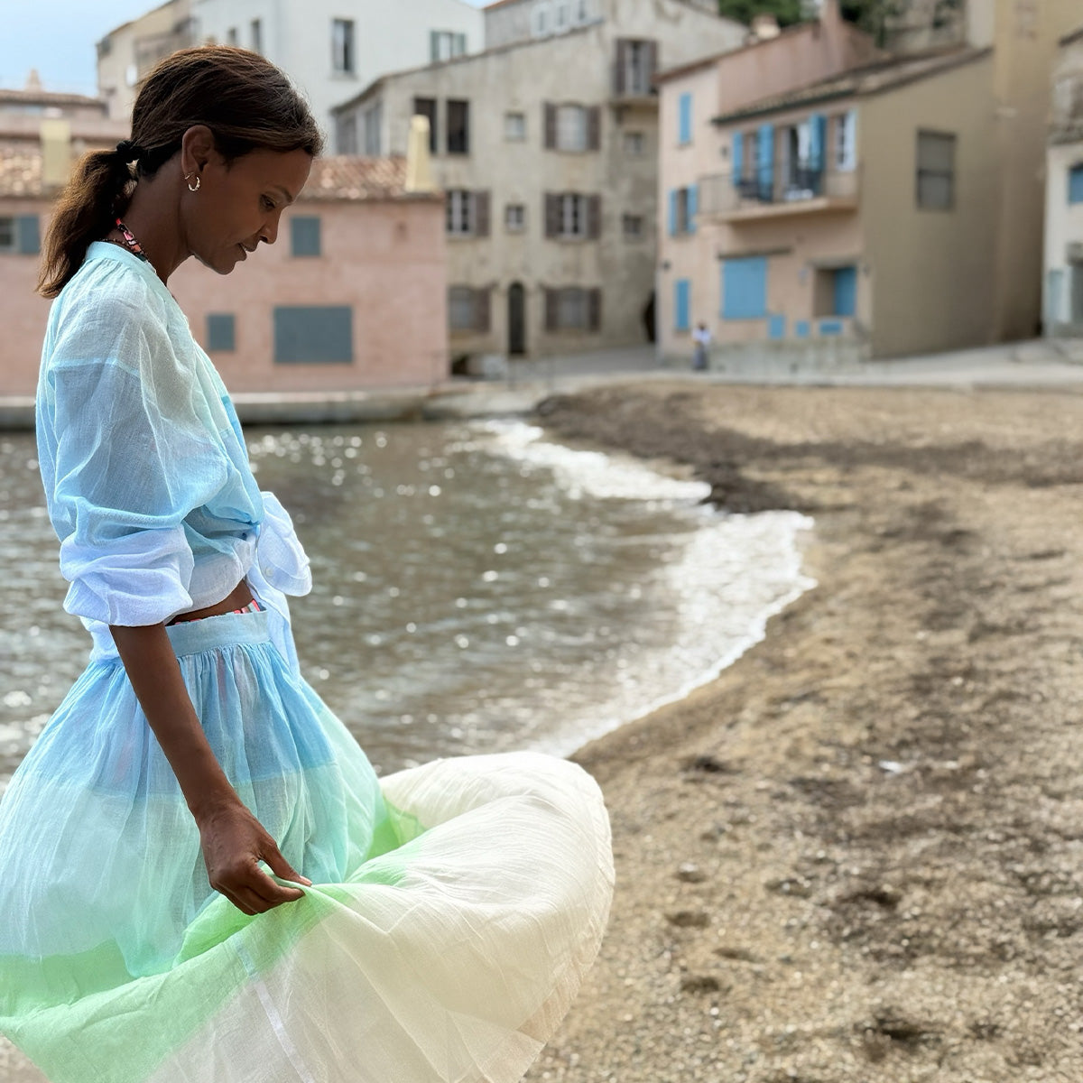 Liya Kebede standing in light handwoveb blue and green skirt standing by a waterfront with a stone wall and building in the background.