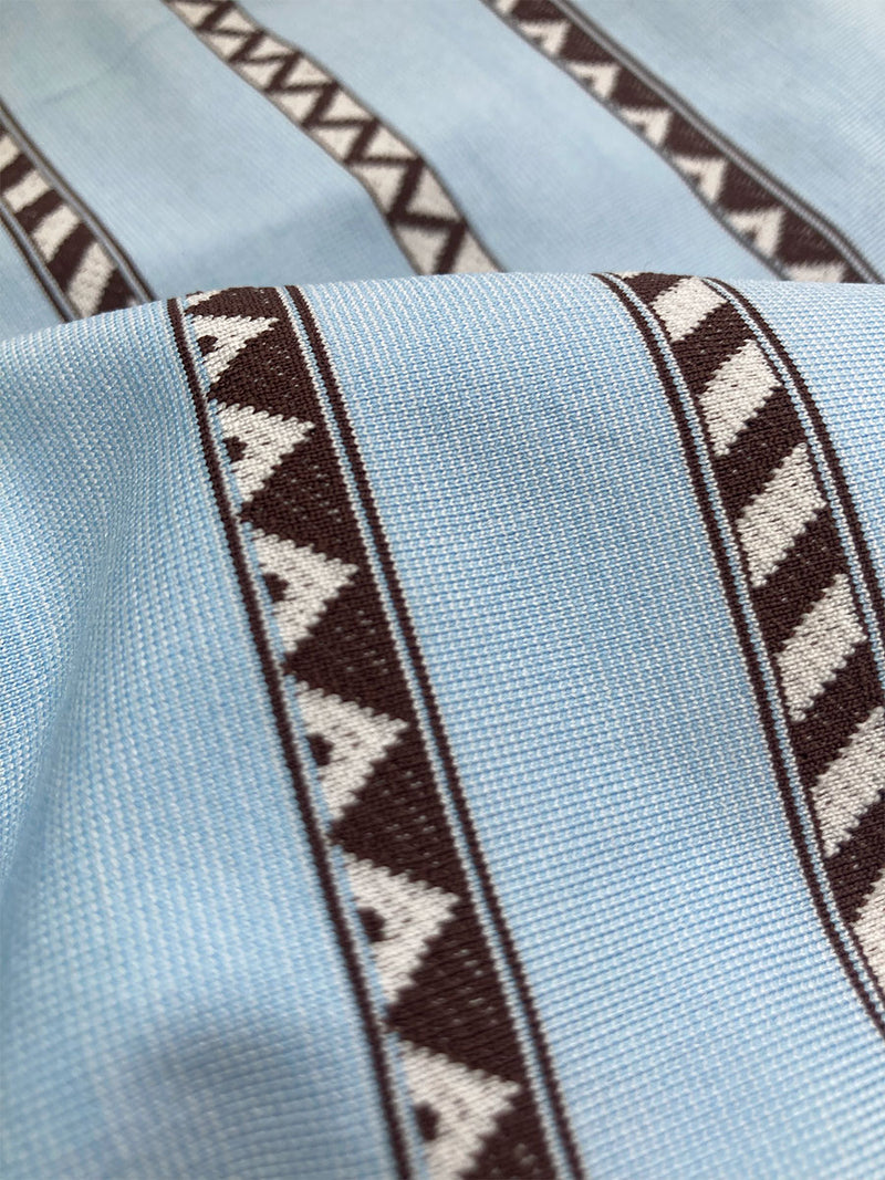 Close up on Tutu Blue Swim Fabric Featuring intricate bands of dark earth and ivory Tibeb patterning on a pale sky blue background.