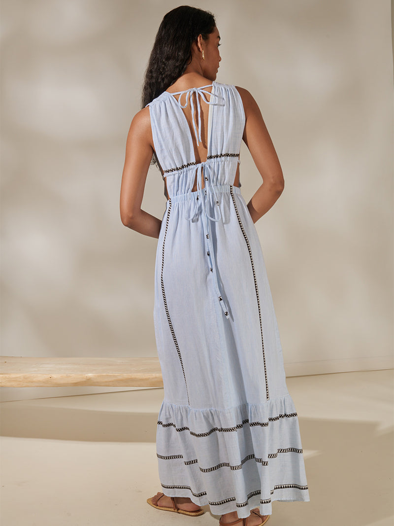 Back view of a woman standing wearing Lelisa V Neck Dress featuring intricate bands of dark earth and ivory Tibeb patterning on a pale sky blue background.