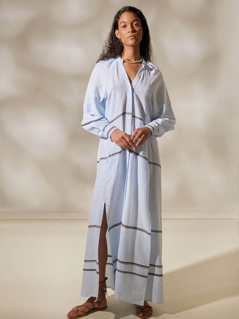 Woman Standing Wearing Makeda Button UP dress featuring intricate bands of dark earth and ivory Tibeb patterning on a pale sky blue background.