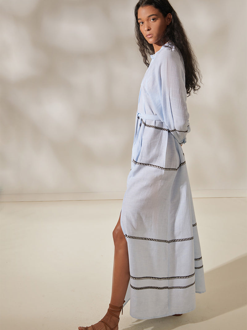 Woman Standing Wearing Makeda Button UP dress featuring intricate bands of dark earth and ivory Tibeb patterning on a pale sky blue background.