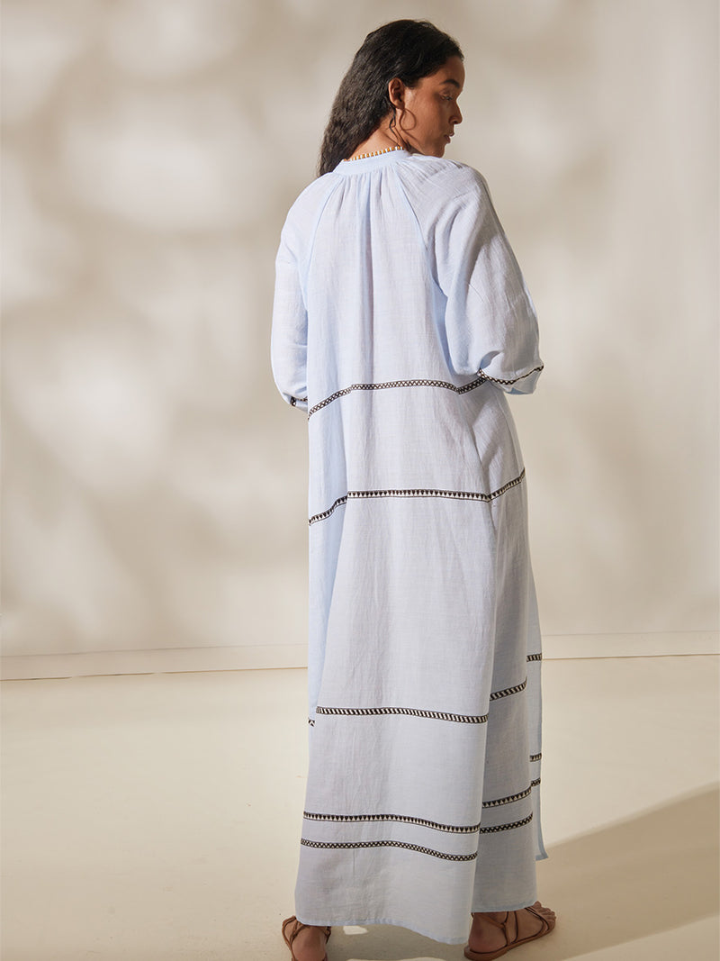 Back of a Woman Standing Wearing Makeda Button UP dress featuring intricate bands of dark earth and ivory Tibeb patterning on a pale sky blue background.