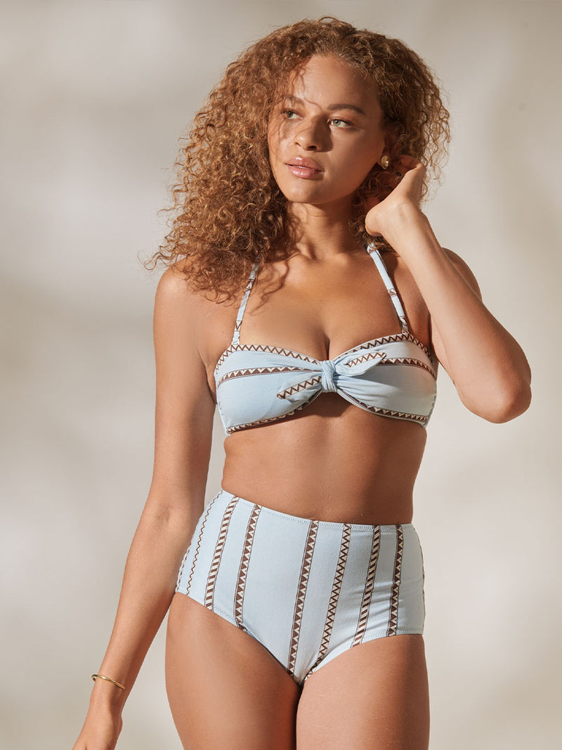 Woman Standing Wearing High Waist Bikini Bottom and AVA Bandeau Top featuring intricate bands of dark earth and ivory Tibeb patterning on a pale sky blue background.