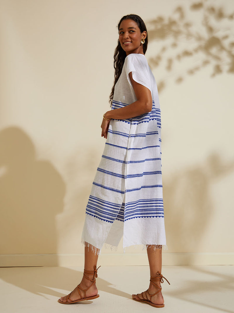 Side view of a woman standing wearing the Yani Caftan Dress featuring blue tibeb diamond design bands on a textured seersucker white background.  