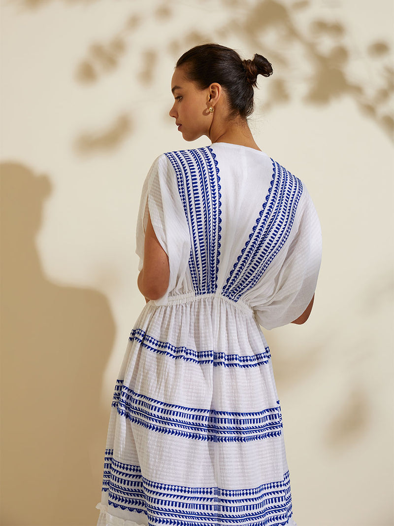 Back view of a woman standing wearing the Yani Plunge Neck Dress featuring blue tibeb diamond design bands on a textured seersucker white background.  