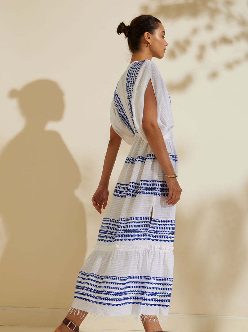 Side view of a woman walking wearing the Yani Plunge Neck Dress featuring blue tibeb diamond design bands on a textured seersucker white background.  