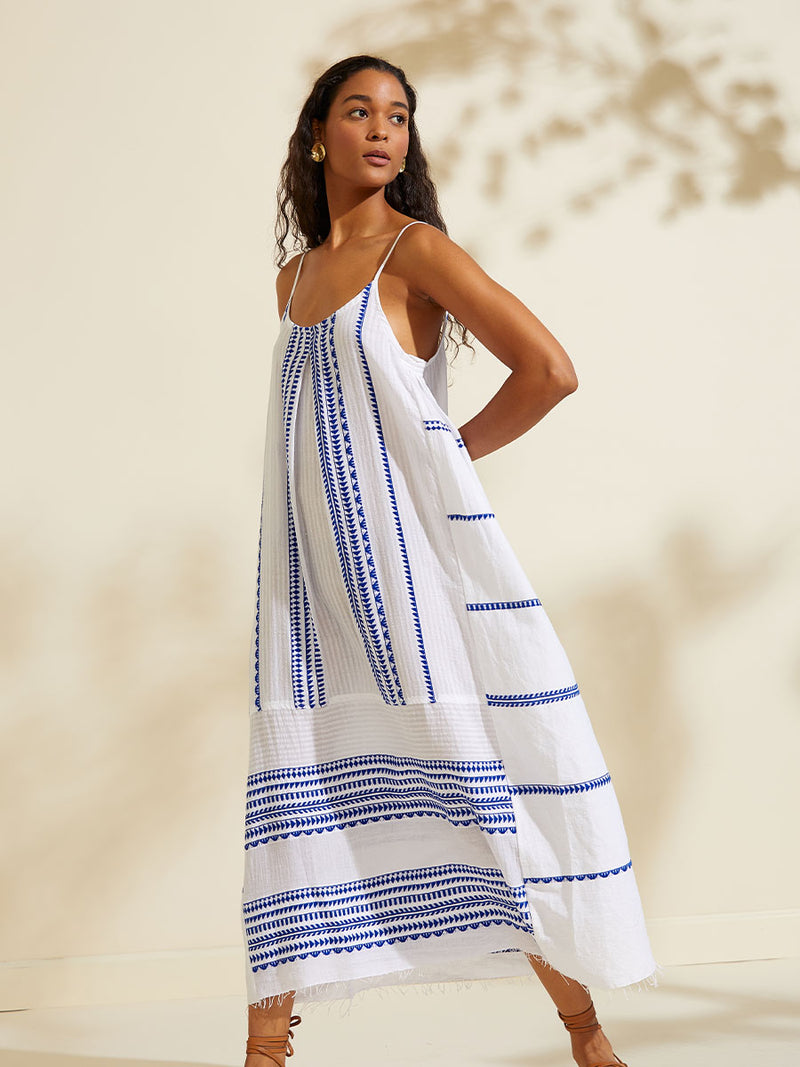 Side view of a woman walking with her arms behind her back  wearing the Yani Slip Dress featuring blue tibeb diamond design bands on a textured seersucker white background.  