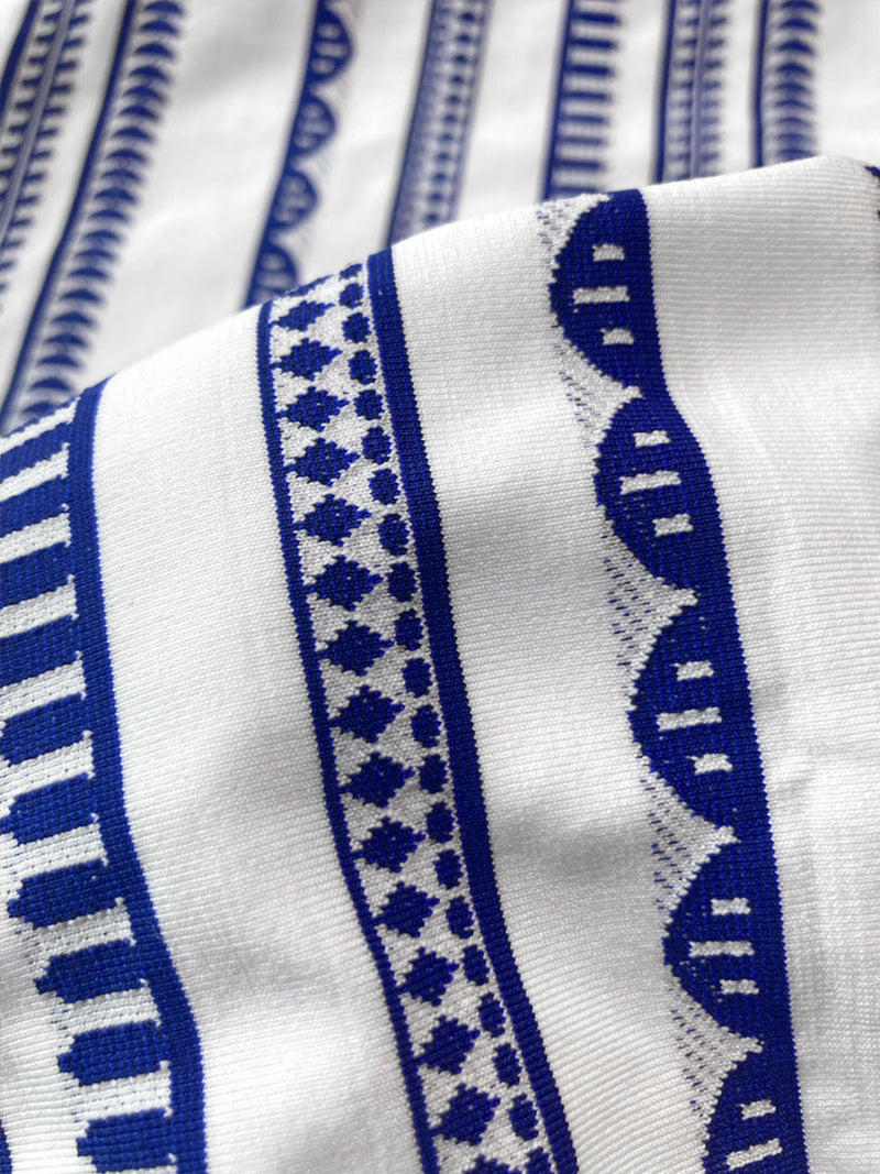 Close up on the fabric of the Yani Side Tie Bikini Bottom featuring blue tibeb diamond design bands on a textured seersucker white background.  
