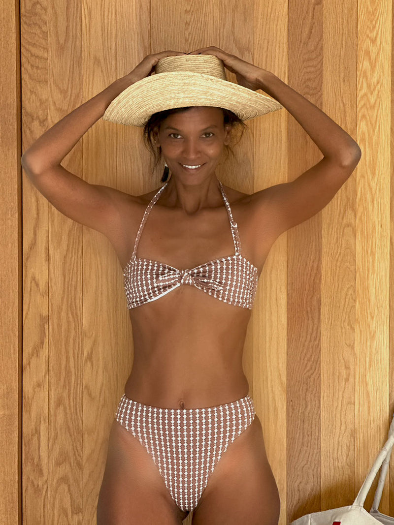 Liya Kebede stands with her arms lifted up, holding a straw hat on her head, wearing a Zala Bandeau Top and Zala High Leg Bikini Bottom