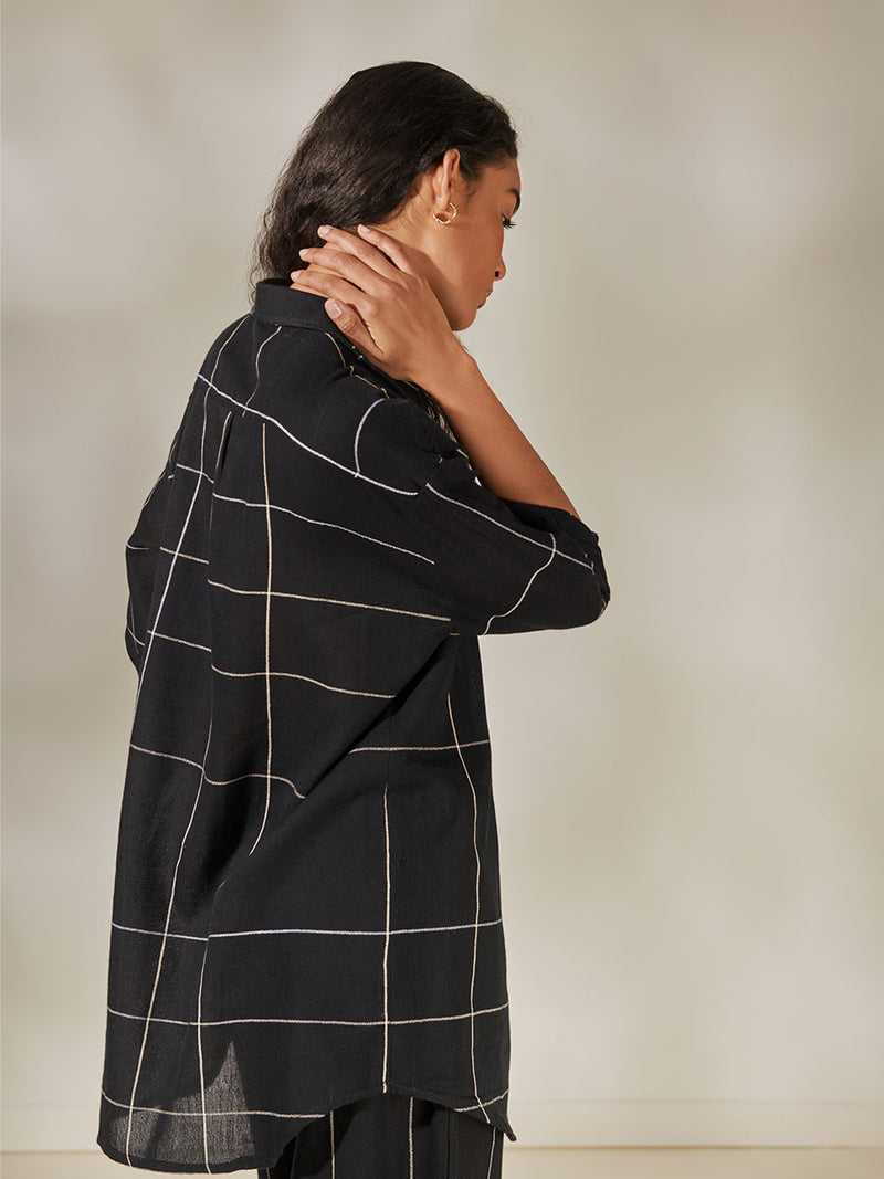 Back Side View of a Woman Standing Wearing Mariam Oversized Shirt featuring Big White Plaid Patten on Black Cotton Background