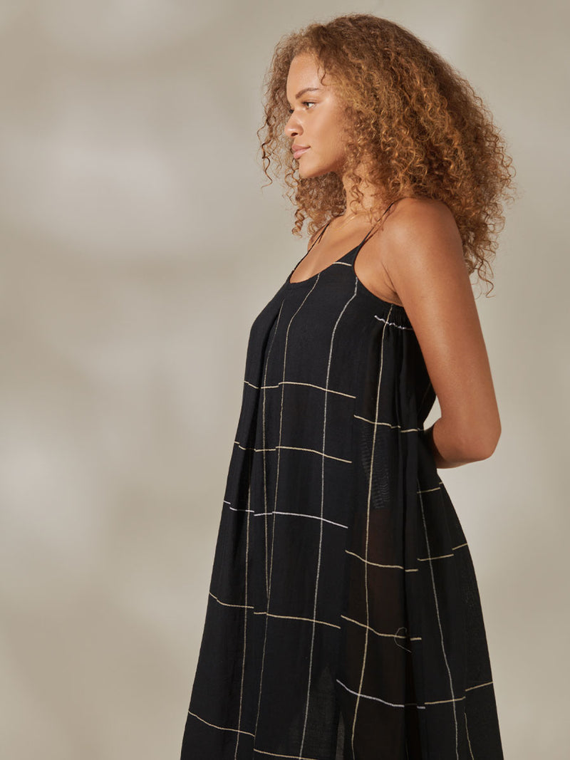 Side View of a Woman standing wearing Nia Maxi Slip Dress featuring Big White Plaid Pattern on Black Cotton Background