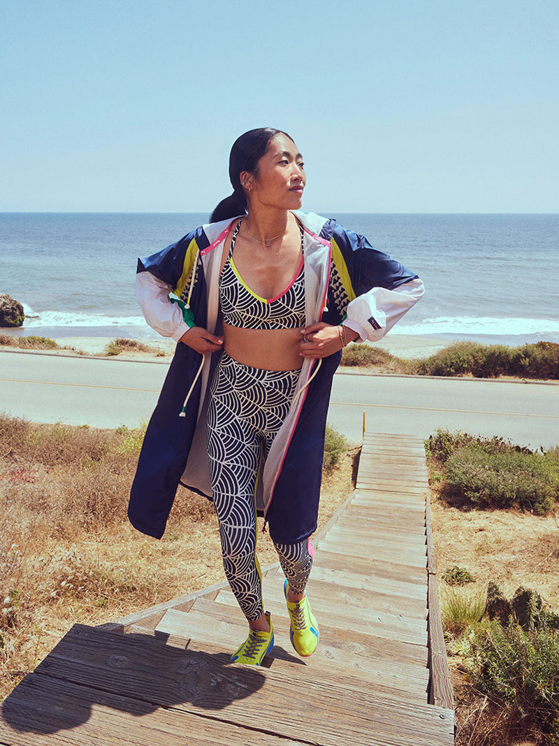 Woman walking on the beach wearing Puma x lemlem Navy Anorak featuring color block details and classic lemlem triangle pattern, low impact bra, hight waist leggings and Yellow Sneakers