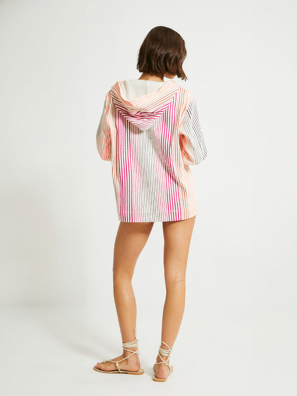 Back of a Woman Standing Wearing lemlem Behati Hoodie Featuring ombre stripe pattern, where deep plum seamlessly transitions into peach, chestnut, and bright pink and lemlem string bikini bottom in magenta color
