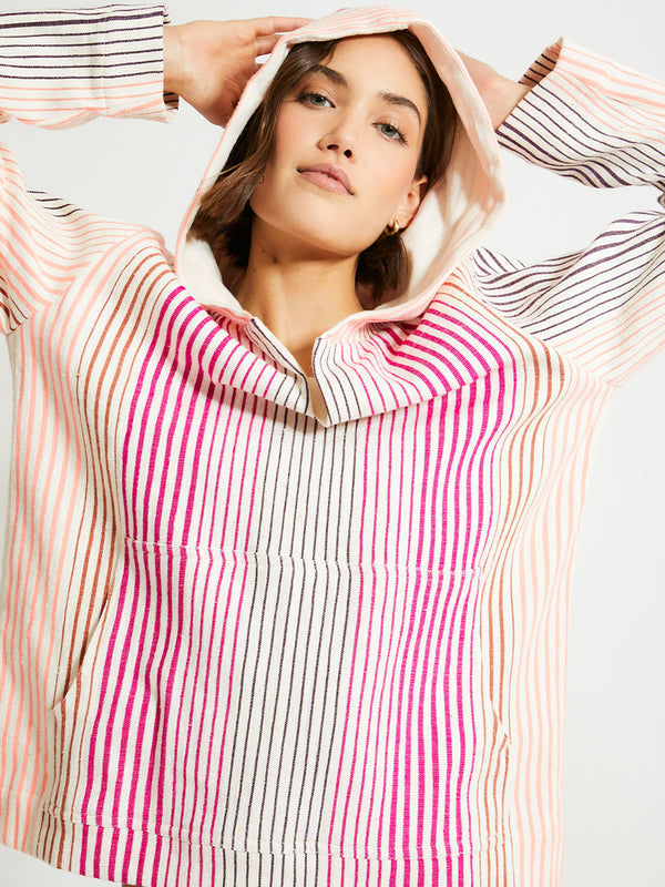 Close up on a Woman Standing Wearing lemlem Behati Hoodie Featuring ombre stripe pattern, where deep plum seamlessly transitions into peach, chestnut, and bright pink and lemlem string bikini bottom in magenta color