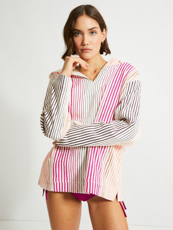 Woman Standing Wearing lemlem Behati Hoodie Featuring ombre stripe pattern, where deep plum seamlessly transitions into peach, chestnut, and bright pink and lemlem string bikini bottom in magenta color