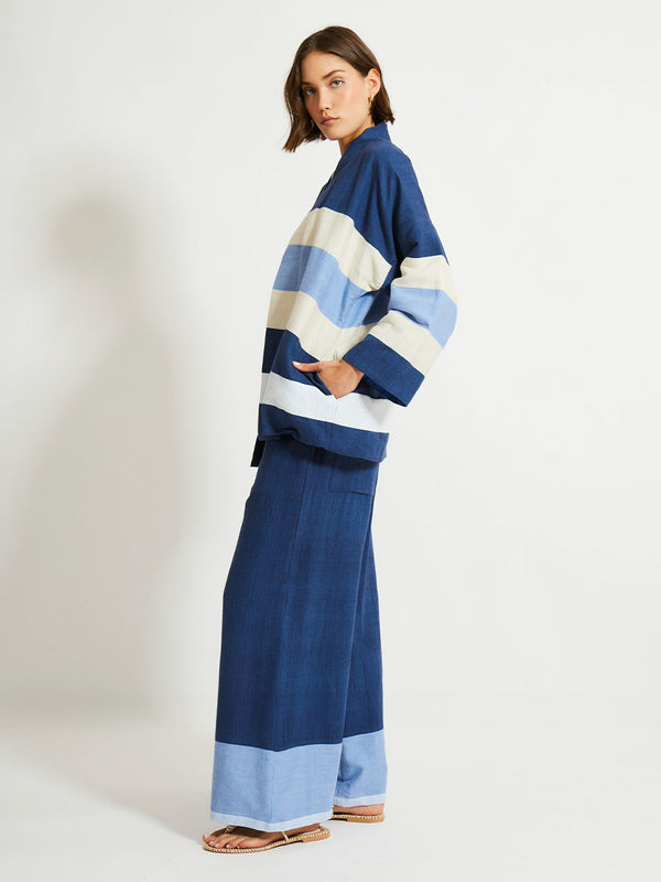 Side of a Woman Standing Wearing lemlem Desta Pants, Terrara Jacket and blouse featuring a striking color-block combination of three shades of blue, paired with bands of neutral cream and white, inspired by handmade indigo quilting and African textil.