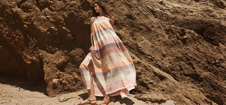 Woman Standing wearing lemlem Amaresh Sunrise Slip Dress featuring stripe pattern in brown, orange, yellow and white colors