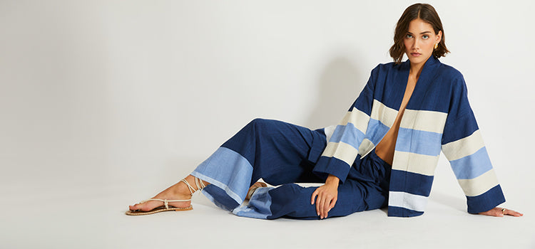 Woman Sitting wearing lemlem Jacket featuring color block pattern in blue. cream and white colors and pants in dark blue color with pale blue accents
