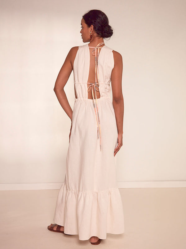 Back View of a Woman Standing Wearing lemlem Lelisa V Neck Dress featuring cream linen tencel accented with vibrant lemlem stripe pattern in warm orange, yellow and peach tones