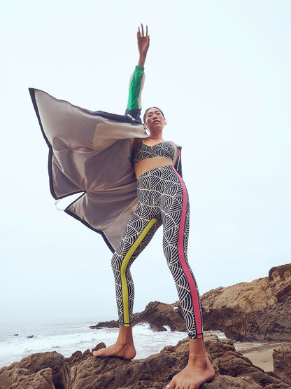 Woman Exercising on the beach wearing Puma x lemlem Leggings in Ghost Pepper and Black colors and matching Puma x lemlem low impact bra featuring hand sketched scallop print and color block accents in bright pink and green colors and Navy Anorak