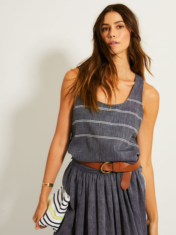 Woman Standing Wearing lemlem Tank Top and a skirt featuring navy color and whote stitching and holding lemlem pouch featuring stripe pattern in black color on white background