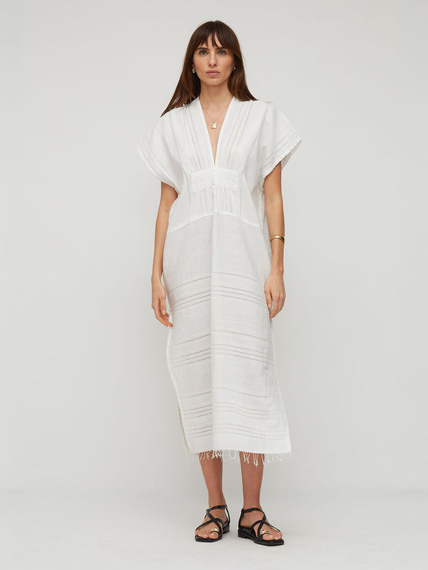 A woman modeling a white lemlem handwoven GASIRA  caftan featuring short sleeves, a deep V-neckline, and horizontal stitched details, paired with black strappy sandals against a light gray studio background.