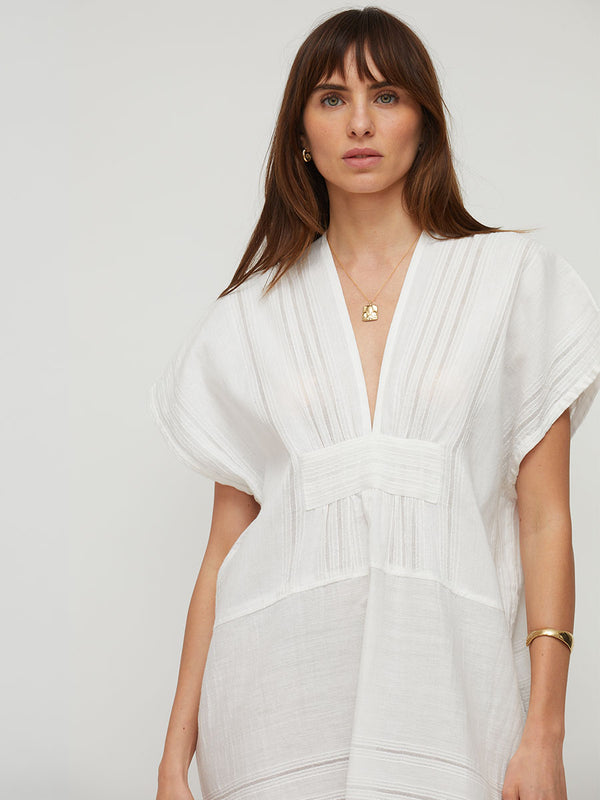 A woman wearing a white short-sleeve lemlem handwoven GASIRA  caftan with a deep V-neck, pleated details, and a gathered waist, photographed against a light gray studio background.